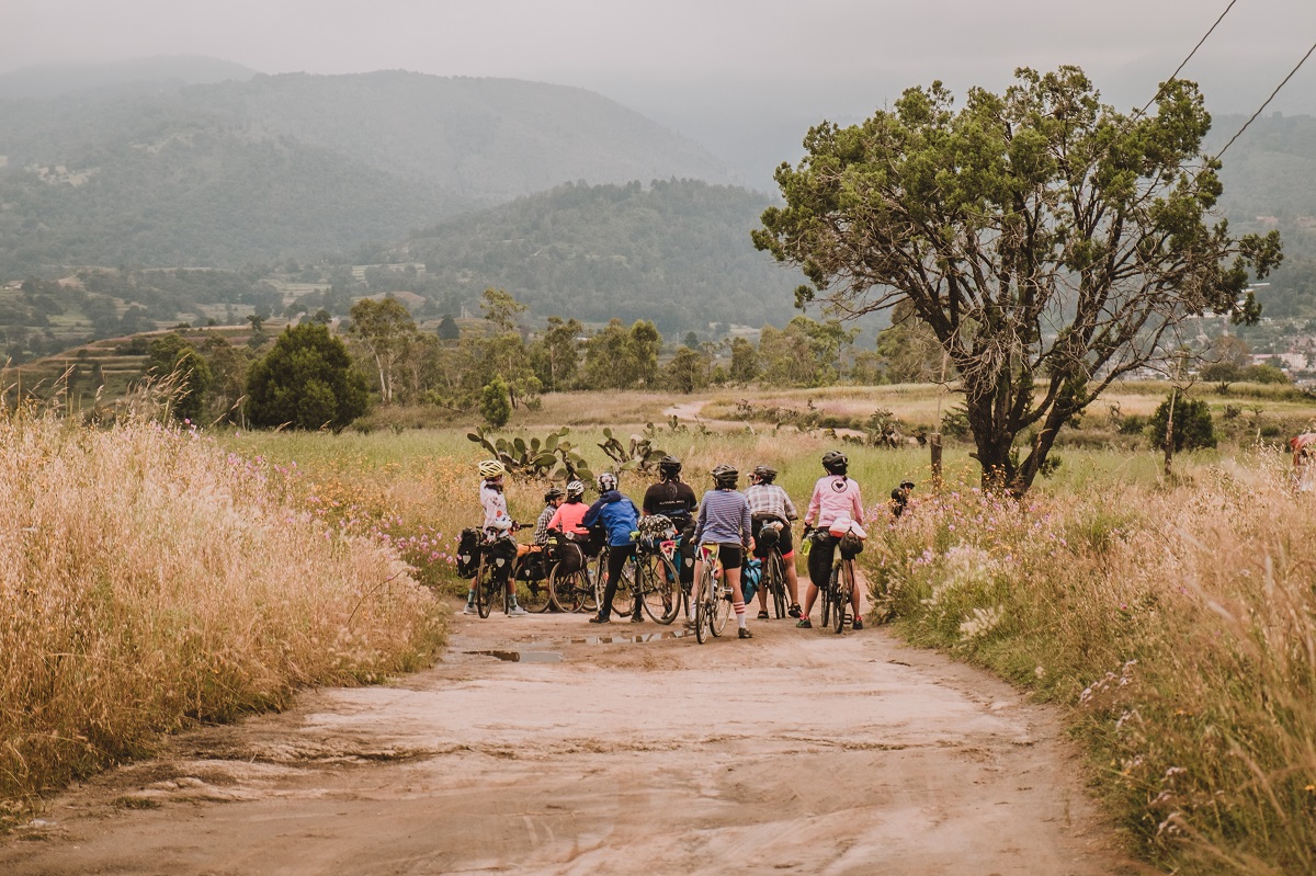 The group of riders huddle in the distance at a grassy dirt road crossing, with a beautiful scene of a rural landscape around them. The green hills in the background are shrouded in mist.