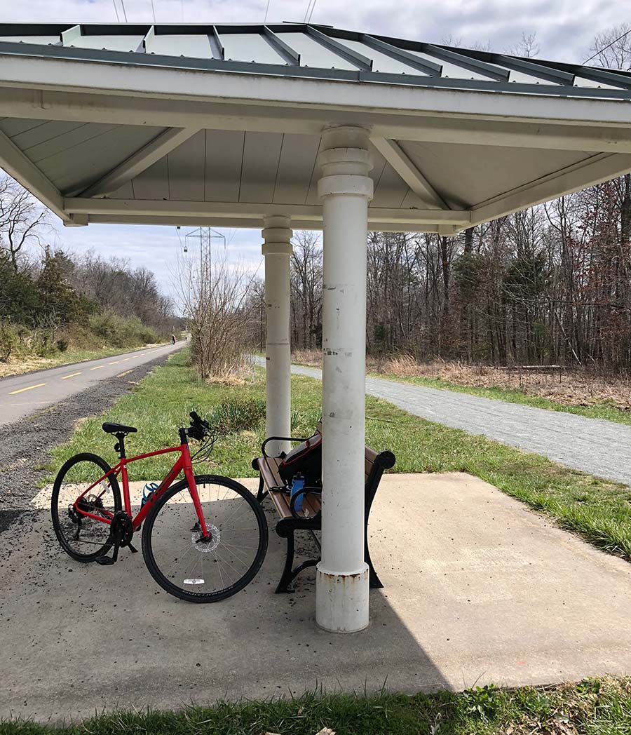Marissa's red bike stands next to the canopied bench along the paved bike trail lined with green trees.
