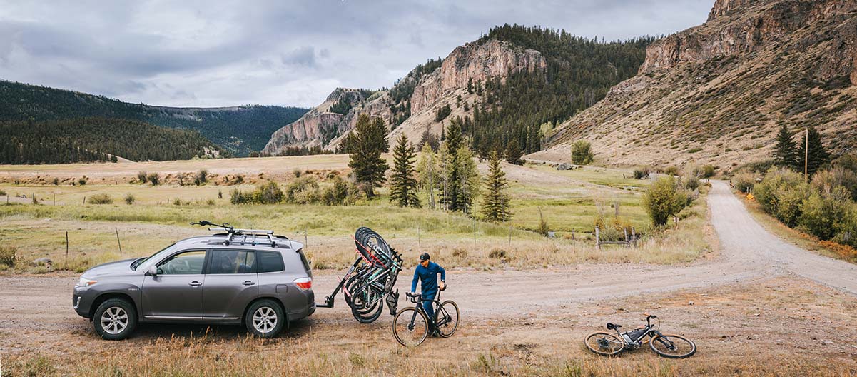 A man loads 6 bikes onto a car's bike rack while parked on a gravel road in the rocky mountains.