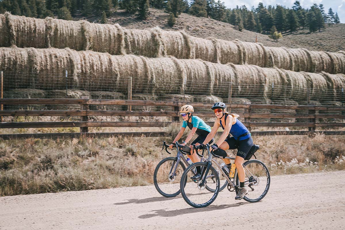 Laura rides beside another woman on a dirt road with haybales behind.