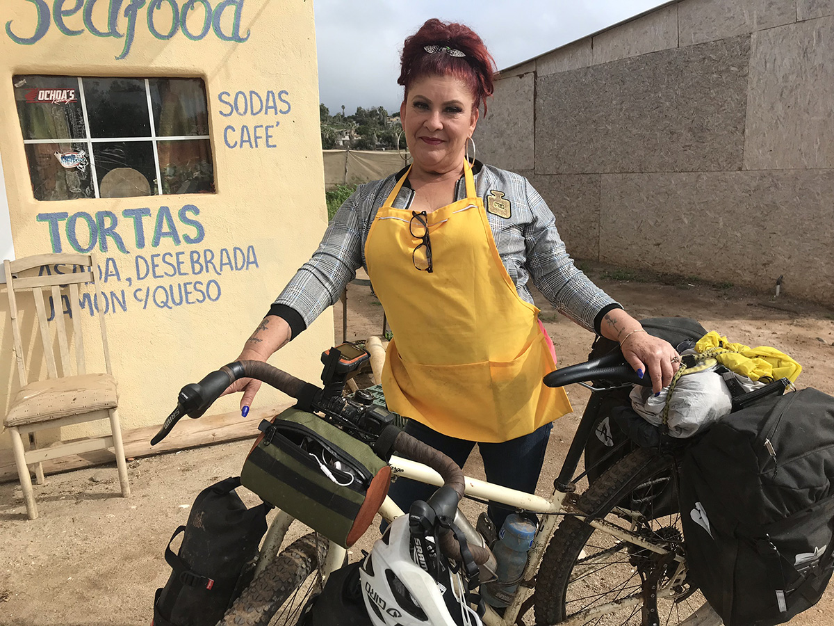 A woman in an yellow apron holds a well-loaded bicycle outside a stucco cafe 