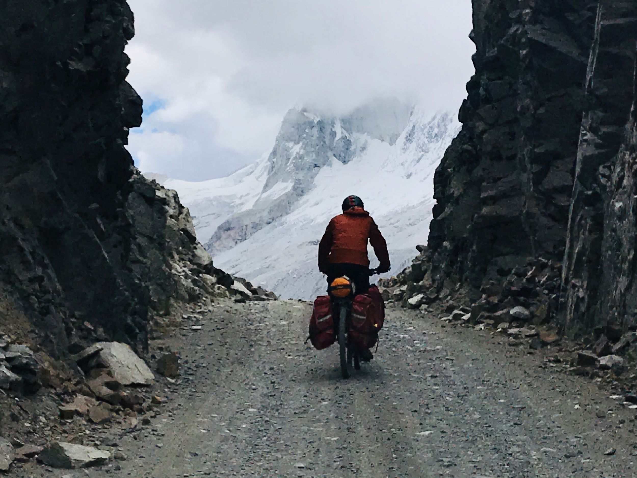 A person rides a loaded bike through a narrow passage between cliffs. Glacier in background.