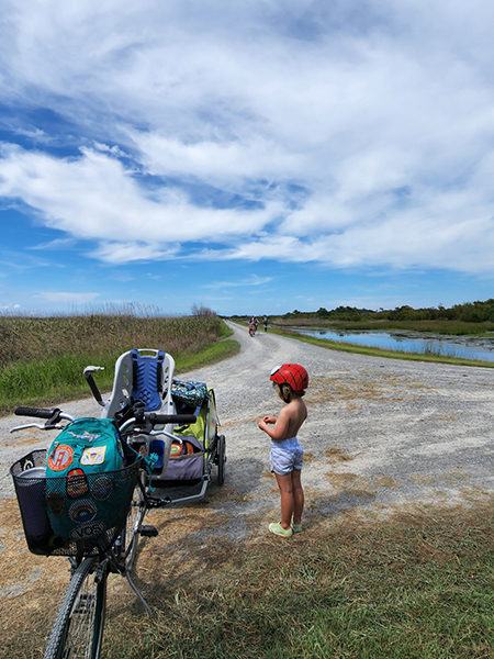 A child stands in the sun next to a bike with a child's seat and trailer on gravel near marshy water.