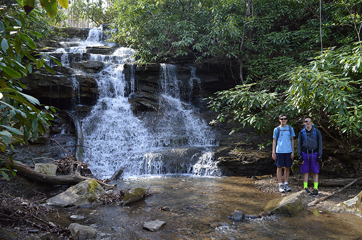 Teenaged boys in shorts stand with their bikes in front of a waterfall.