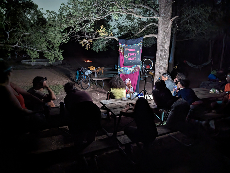 A child sings into a microphone, illuminated by a bright light, with the Bike Story Night banner hanging from a tree.