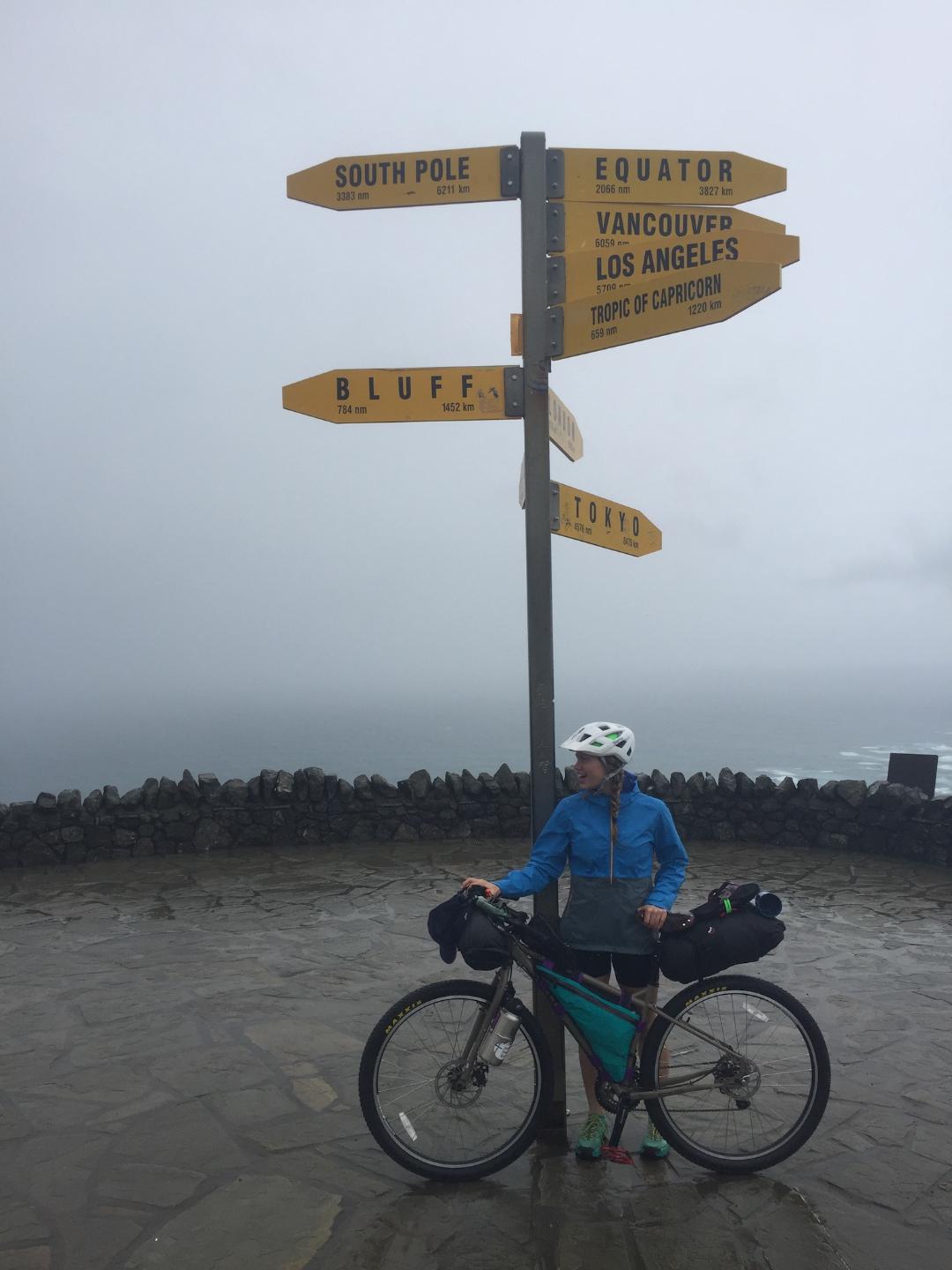 A woman stands with her loaded bicycle underneath signs pointing to important places in the world on a wet, misty coast.