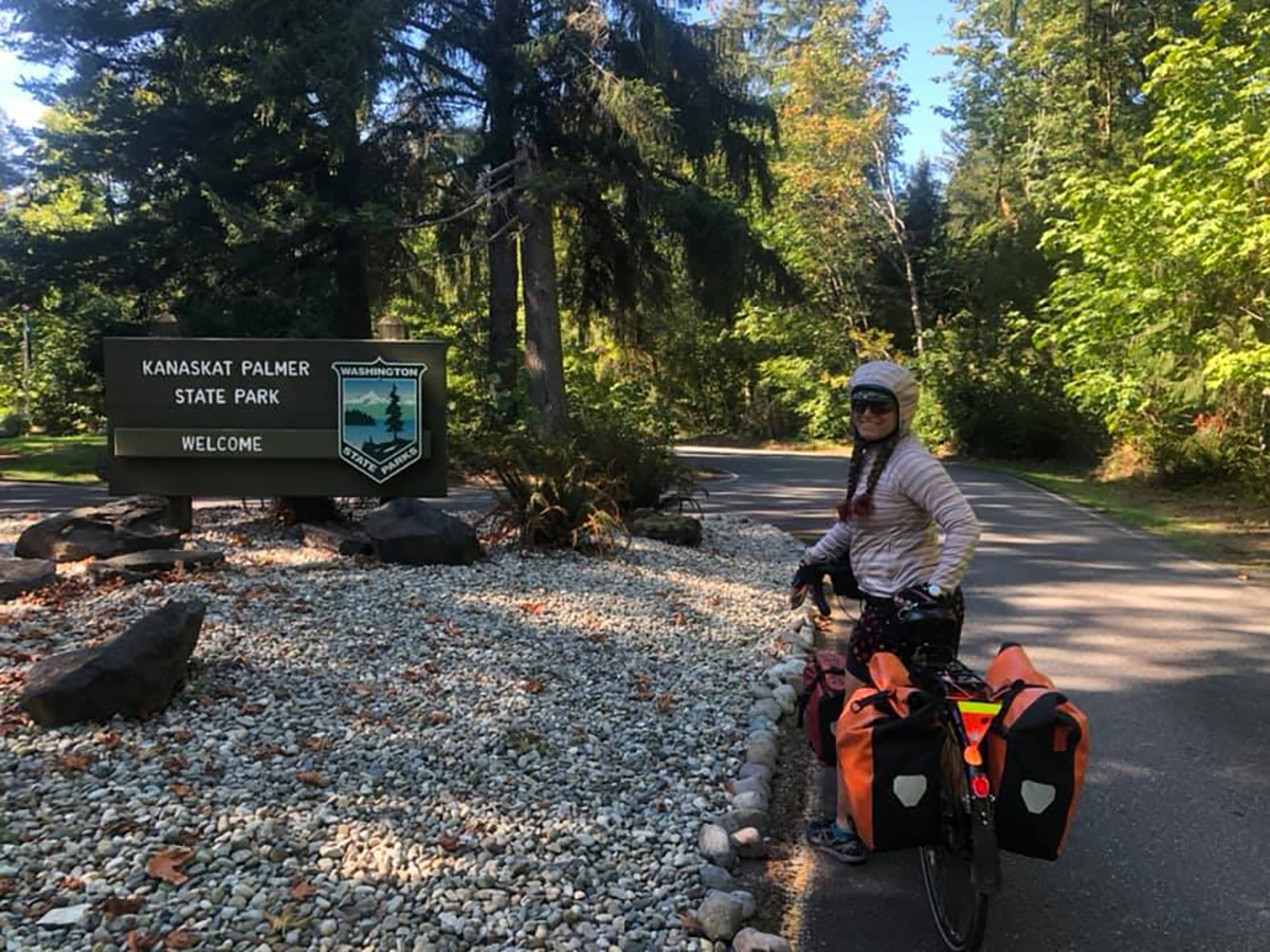 A woman smiles straddling her loaded bicycle in front of a sign: Kanaskat Palmer State Park. Welcome.