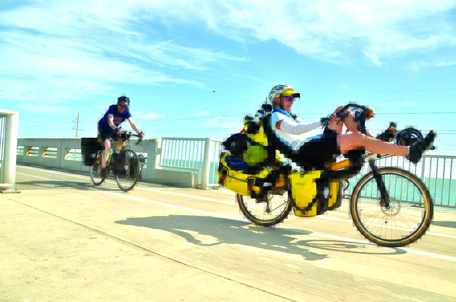 A man in a recumbent cycle and a man on a standard bicycle smile as they go over a bridge.
