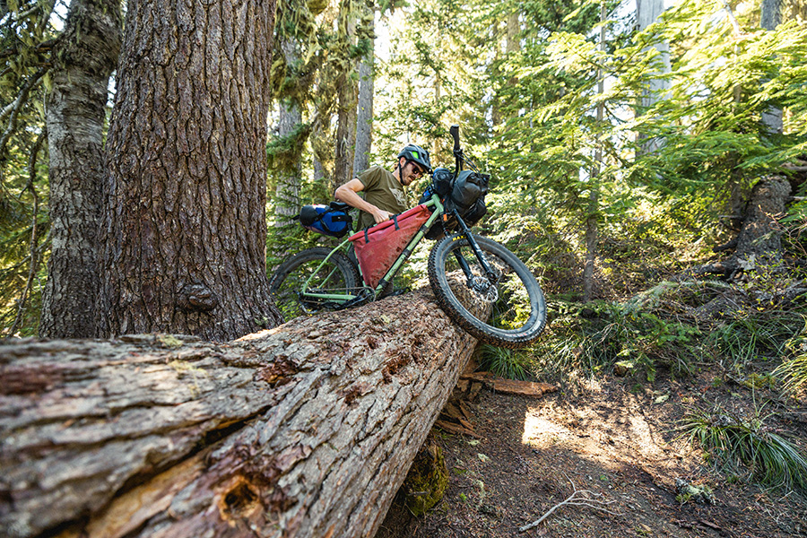 A man lifts his loaded bike over a large log.