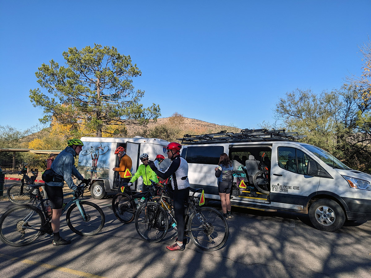 Cyclists retrieve their bikes and gear from a van and listen to instructions.