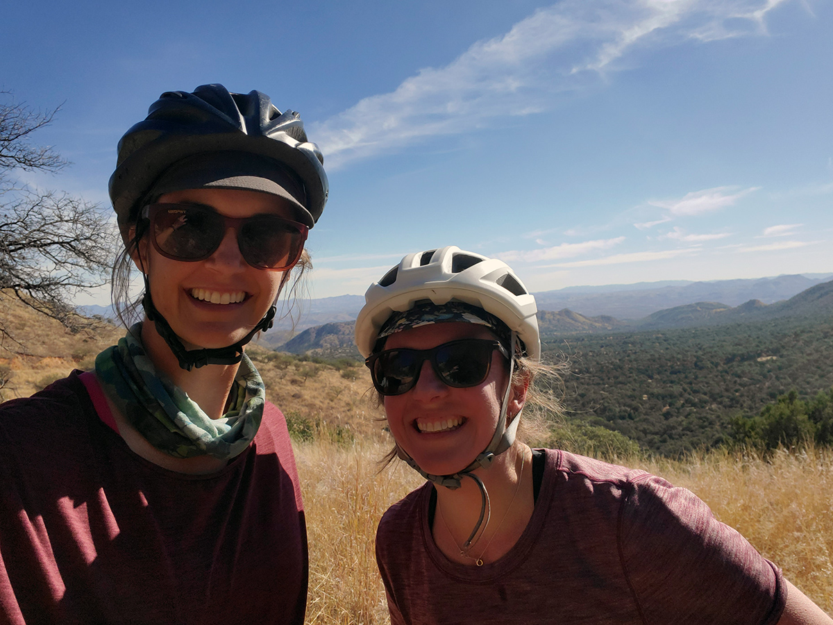 Two women wearing sunglasses and bike helmets smiling for the camera.