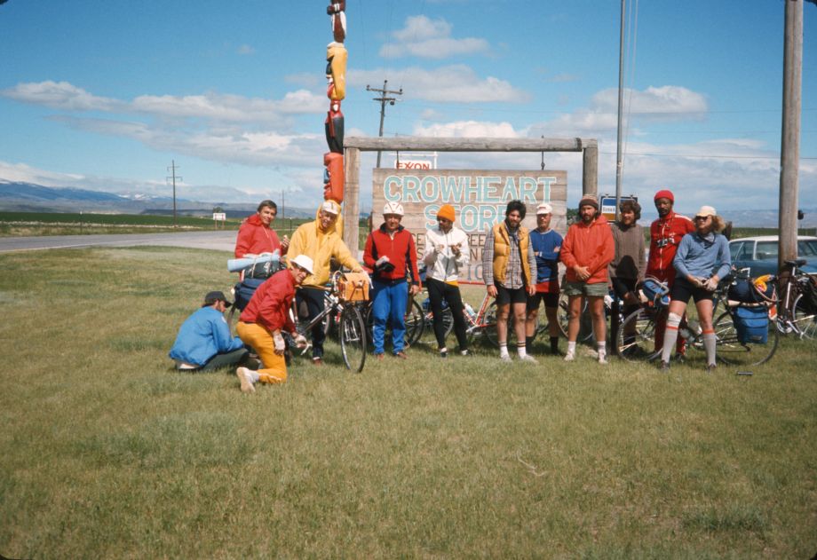 Twelve men on bikes standing on grass in front of a sign for Crowheart, WY