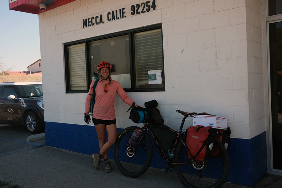 A woman in cycling gear with a loaded bike stands outside the Mecca California post office