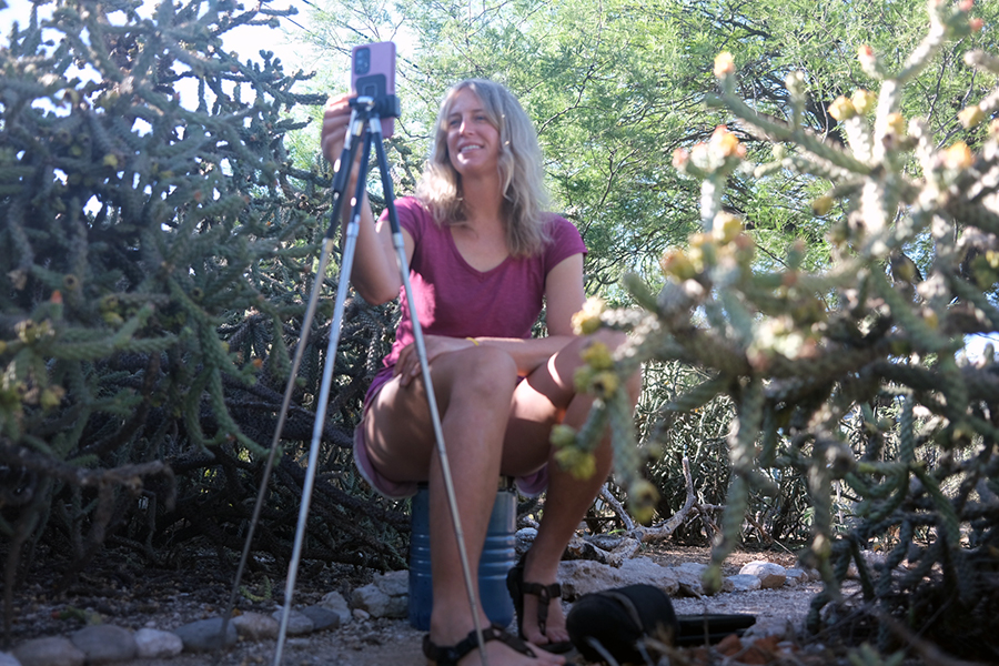 A seated woman smiles for a phone on a tripod in between cactuses.