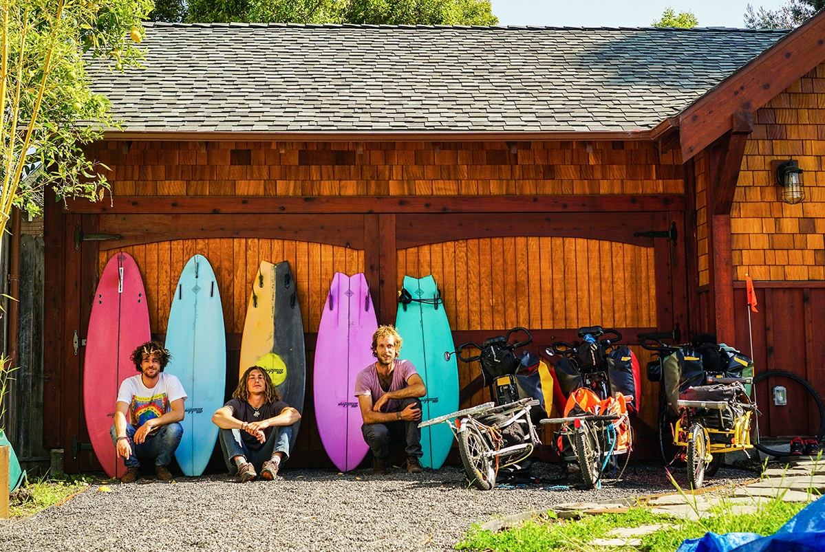 Three men squat against a barn with their loaded bikes and surf boards