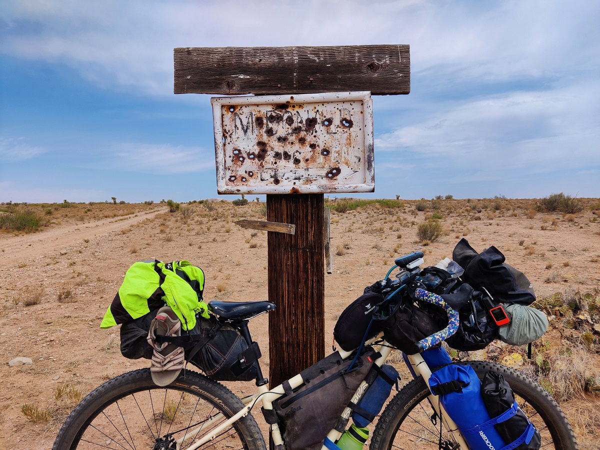 A full loaded, fully rigid bike leans against a sign full of bullet holes. It's unreadable. The landscape is bare and brown and yet still pleasant.