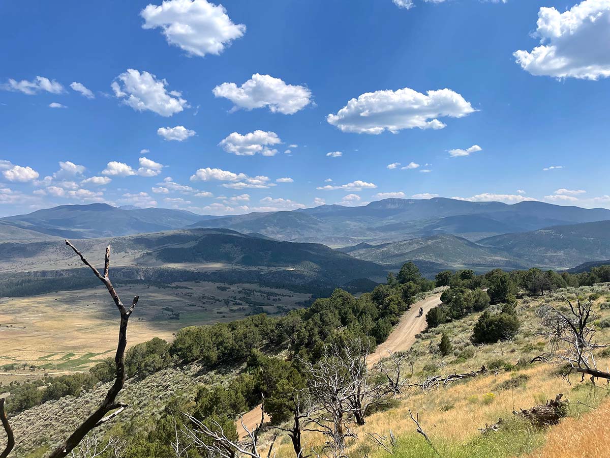 A tiny cyclist in a big, remote landscape of mountains, clouds, blue skies, and one lone dirt road winding among it all.