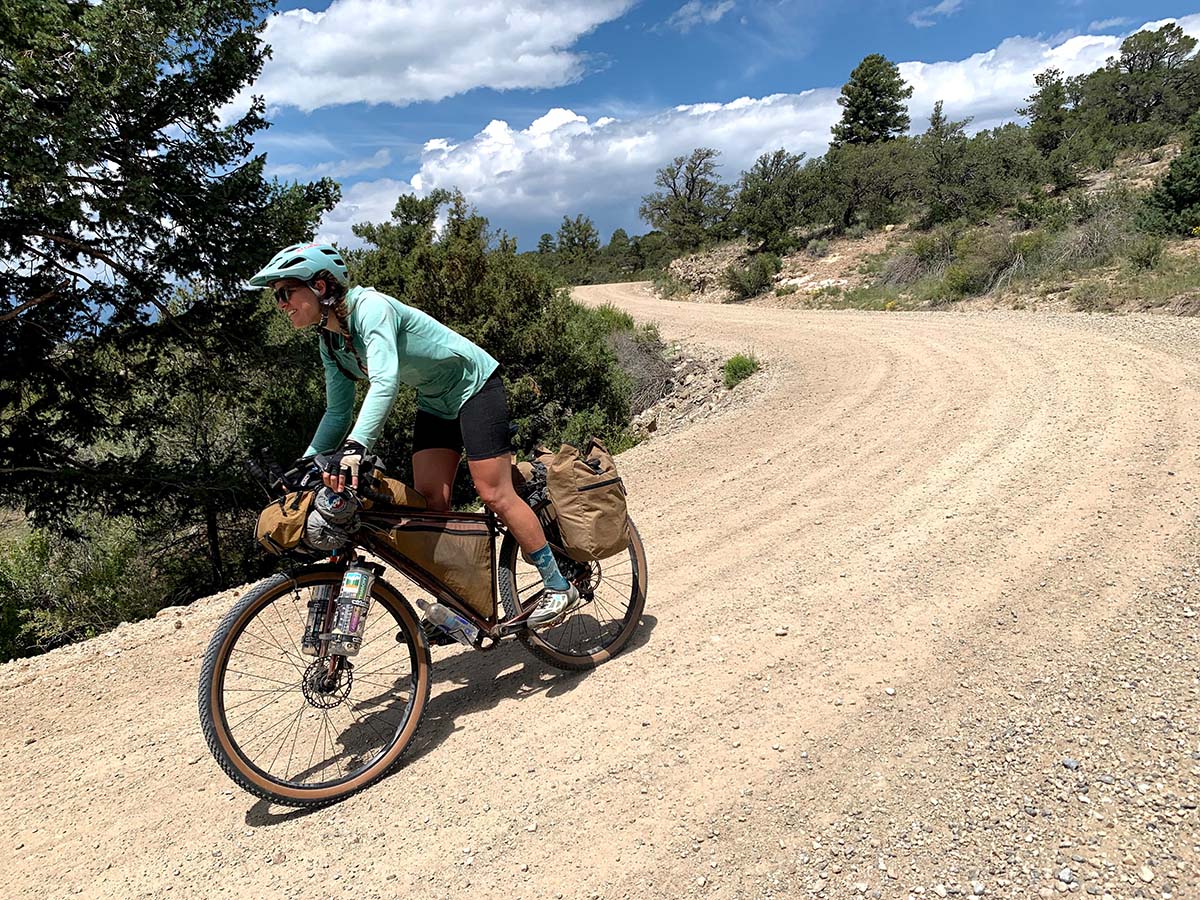 A white woman with brown hair rides her full loaded bike down a steep dirt road.