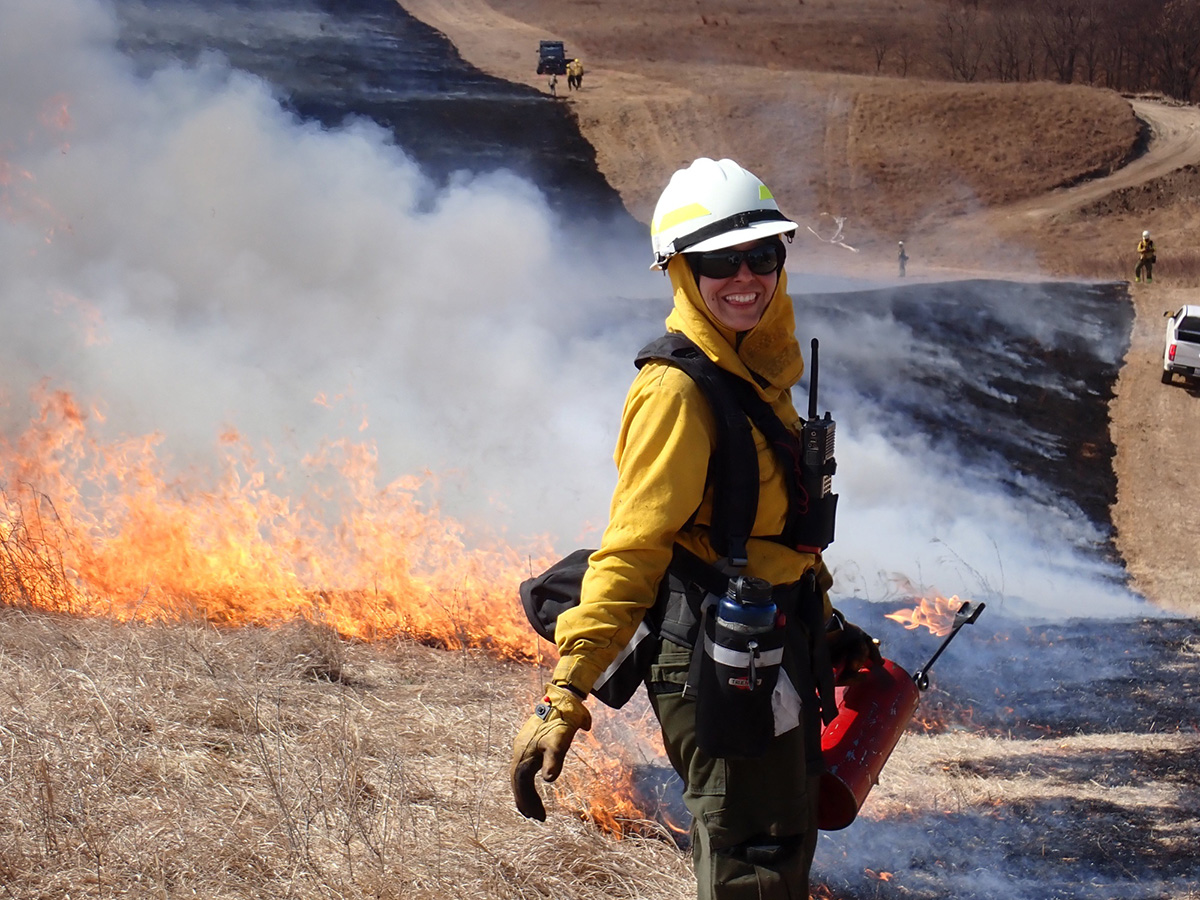 The photo shows a young white woman in heavy work clothes and a helmet holding a blowtorch andsmiling next to a prescribed burn she is a working on.