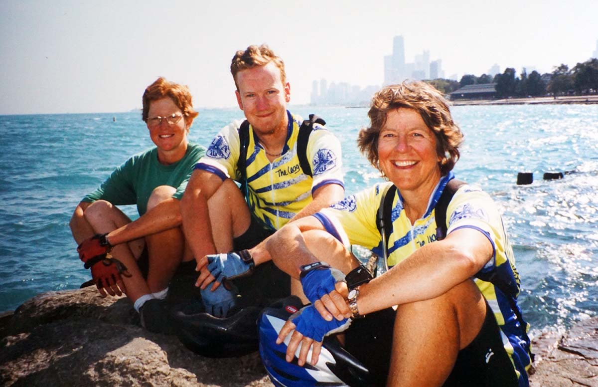 Three white people sit next to Lake Michigan with big flat water and the skyscrapers of Chicago in the background. Two are young and one is middle-aged.