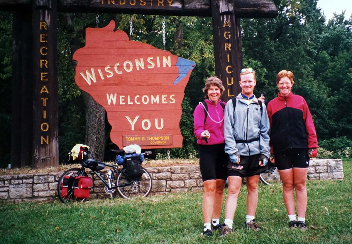 The three riders stand next to a large wooden sign in the shape of Wisconsin saying "Wisconsin Welcomes You".