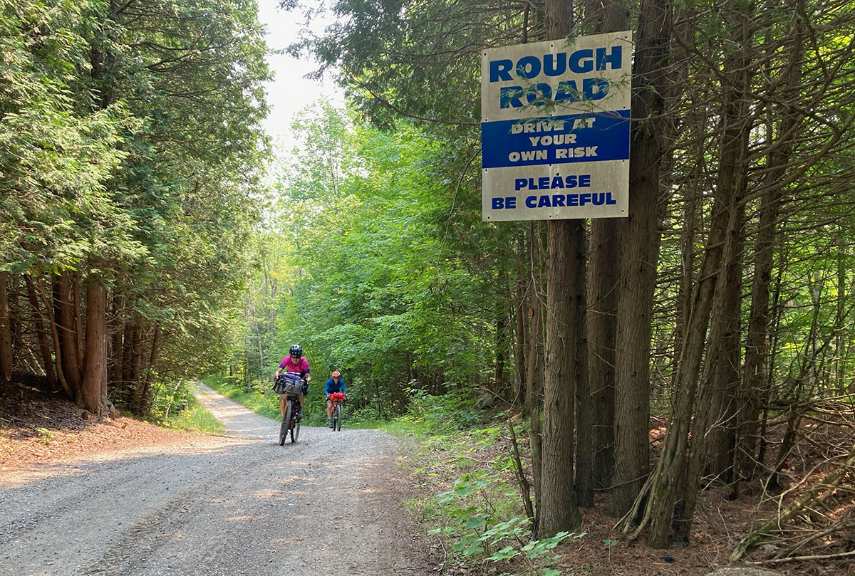 Two cyclists ride on a gravel road toward a sign on a tree that says "Rough Road. Drive at your own risk. Please be careful."