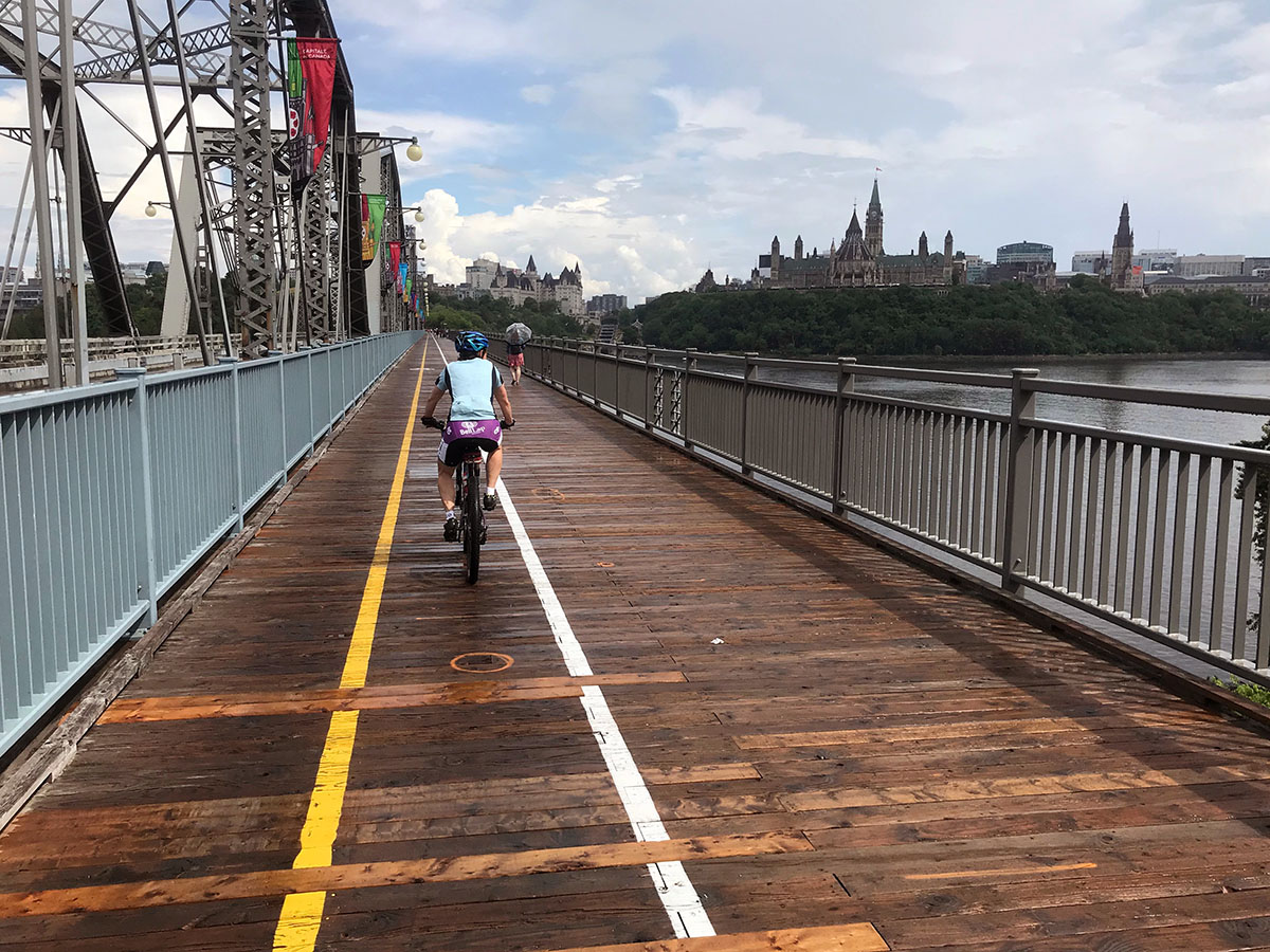 The picture shows a cyclist riding across a wood and metal bridge into Ontario. The rider is on a nice, divided pedestrian part of the bridge. A cityscape and trees are visible on the other side of the river.