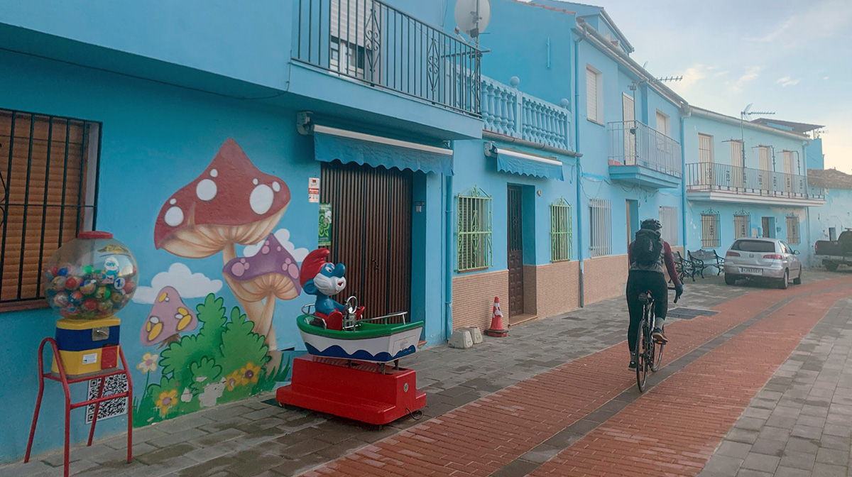 Gina's friend rides along a street in town that is lined with bright blue painted houses and smurf murals.