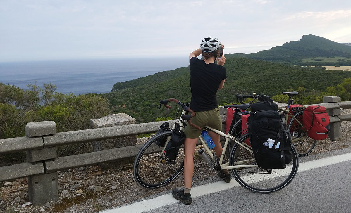 Hollie stands over her bike on the edge of a paved road to take a photo of the coastal view: hills, trees, and ocean.