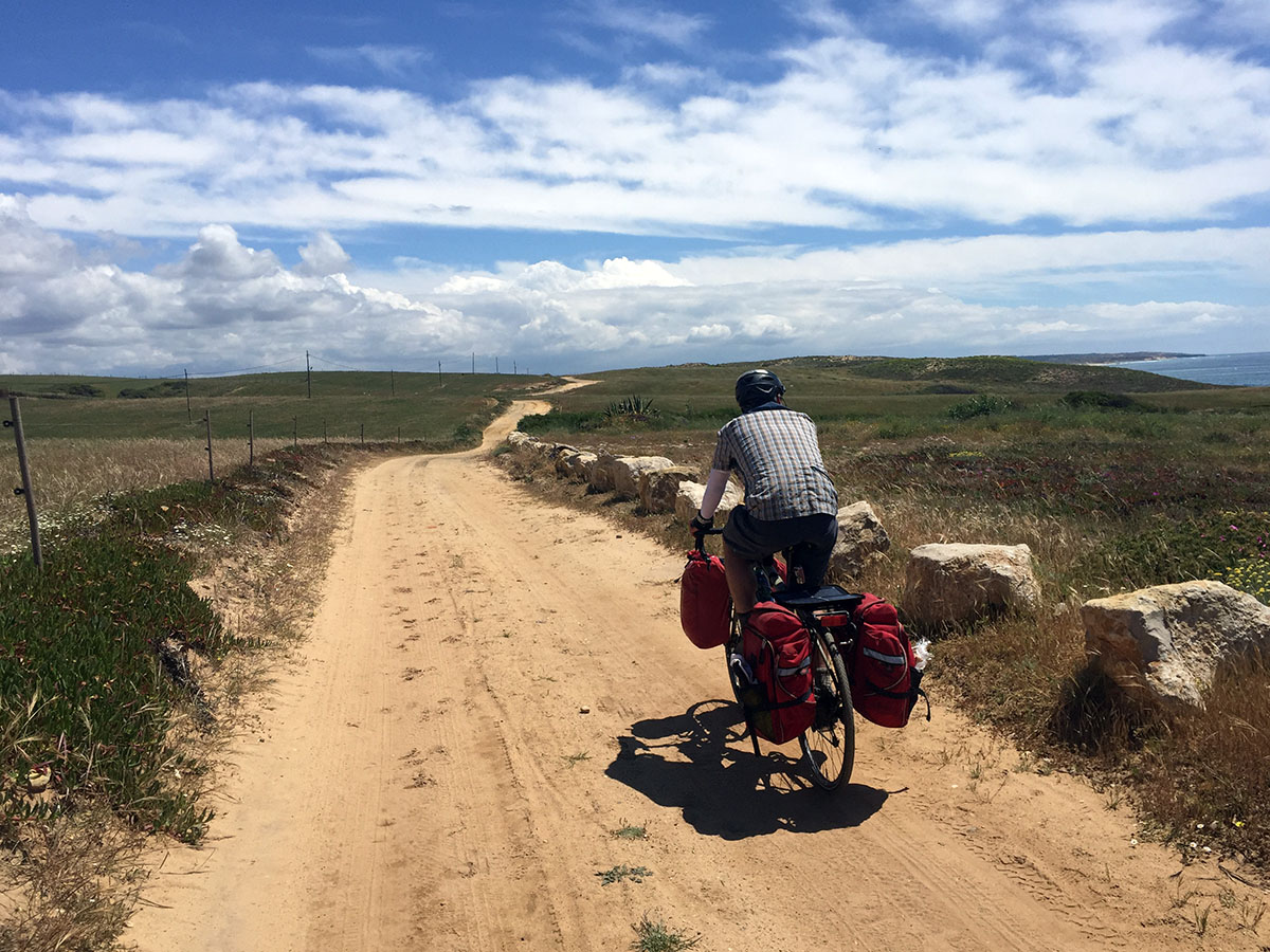 Tom rides on a sandy dirt road near the coastline, ocean in view. There are no trees, just grasses and flowers on a beautifully sunny day.