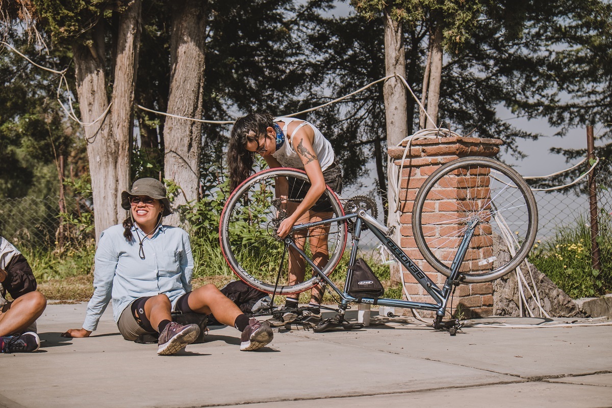 A brown-skinned, dark-haired person lounges on the sidewalk in the sun with their helmet on while the person beside them fixes a flat tire on an upturned bicycle.