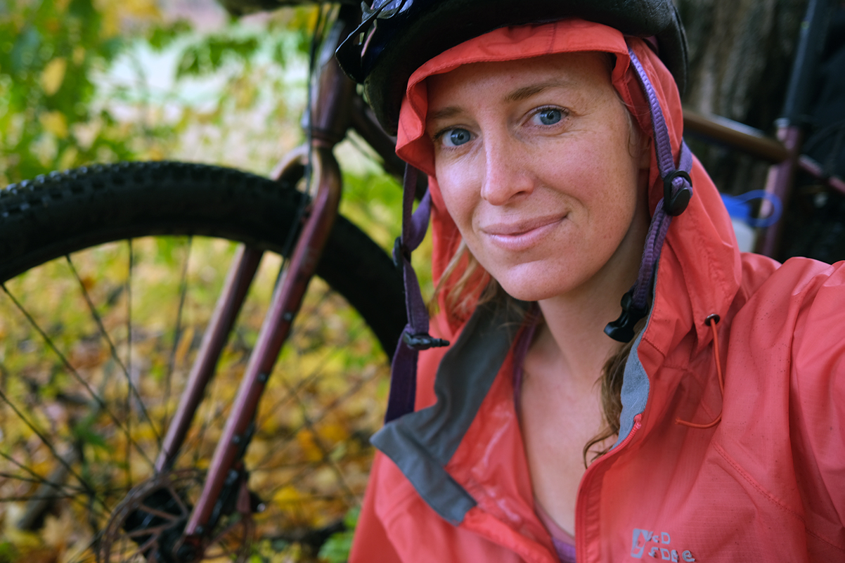 Laura sits in front of her bike covered in rain gear but happy. The autumn leaves make for a colorful backdrop.