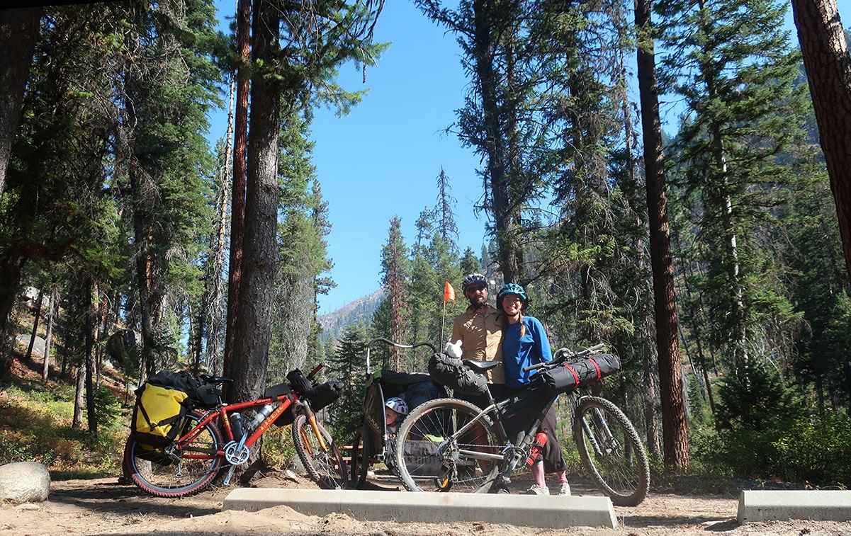 Melissa and her husband stand with their bikes, trailer, and kid in front of a stand of pine trees.