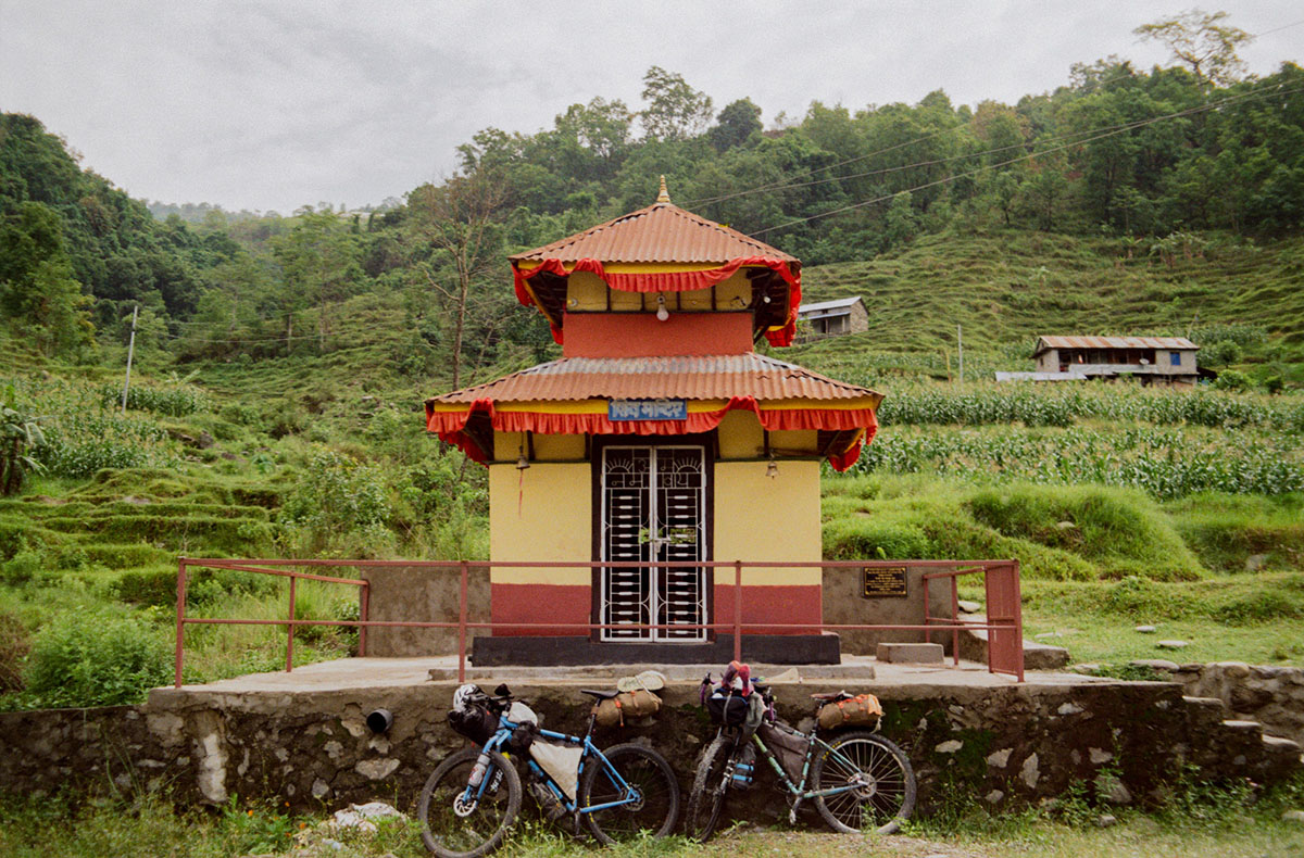 Two bikes lean against the outside railing of small but ornately decorated yellow and red building in a rural and green setting.