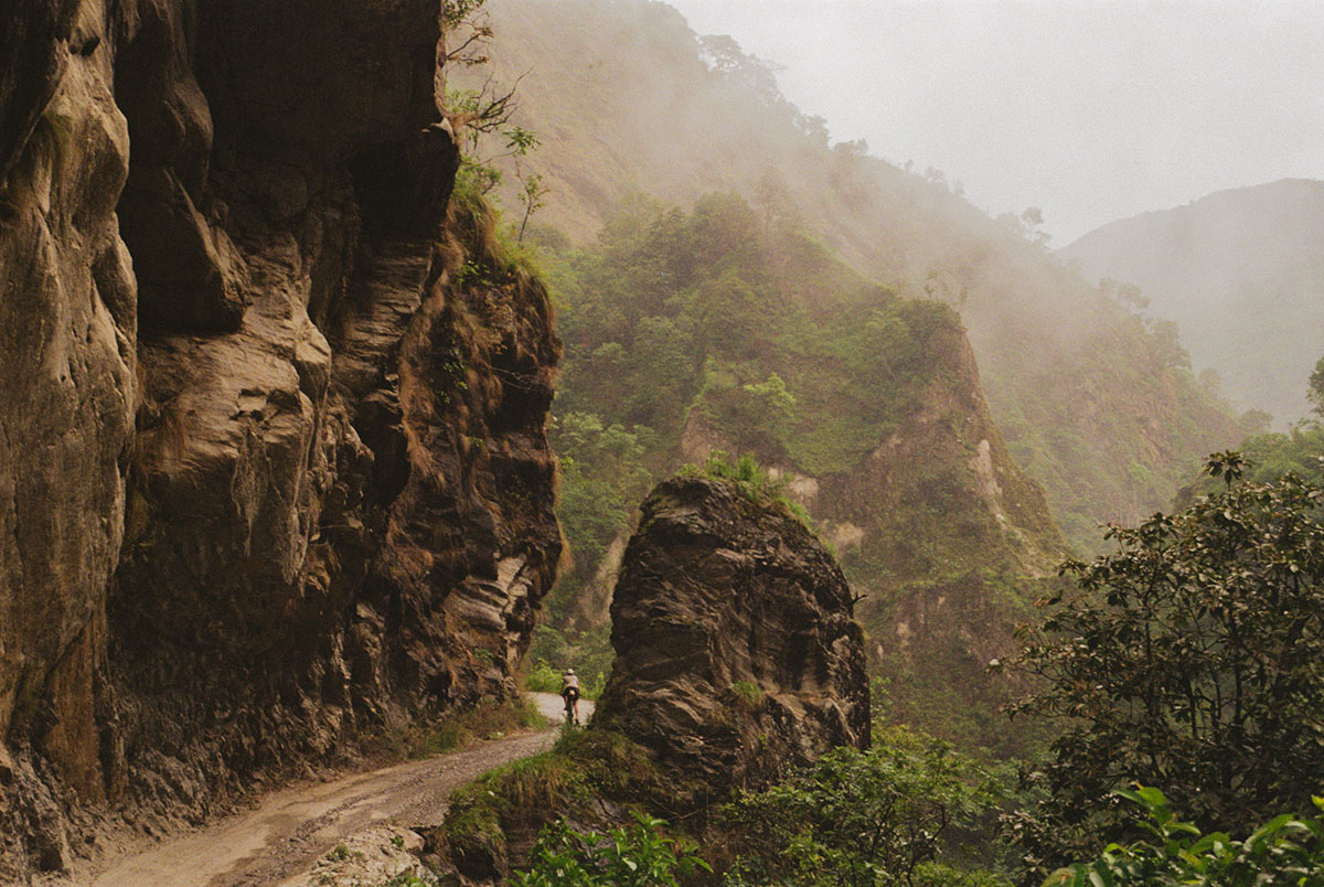 A small cyclist ahead on a dirt road that winds through foggy cliffs and green vegetation.