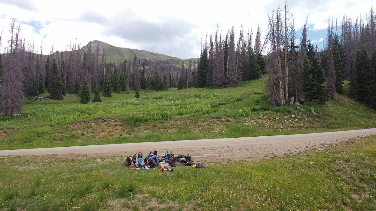 Pak and his two companions, all Korean American men, rest next to a dirt road in a green alpine meadow with few trees.