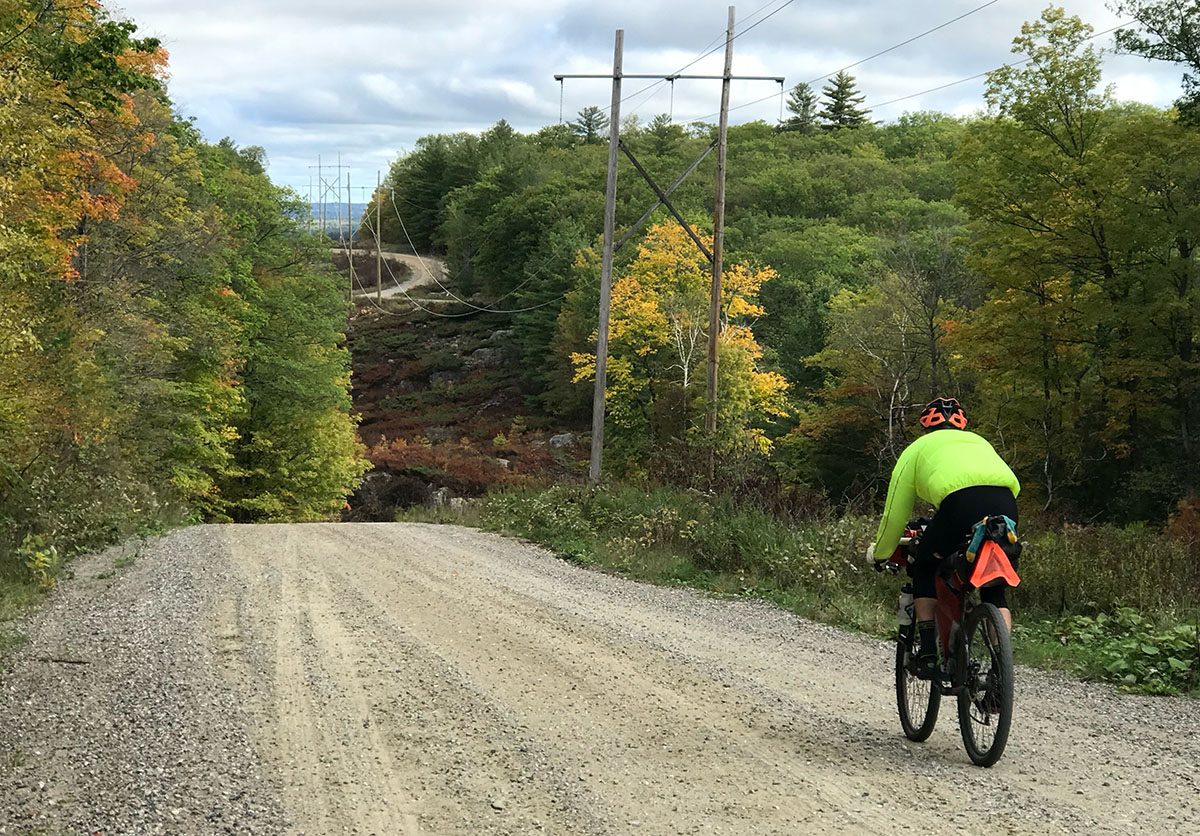 Autumn colors are just beginning to show in the trees and shrubs along a gravel road that a cyclist is riding down a hill.