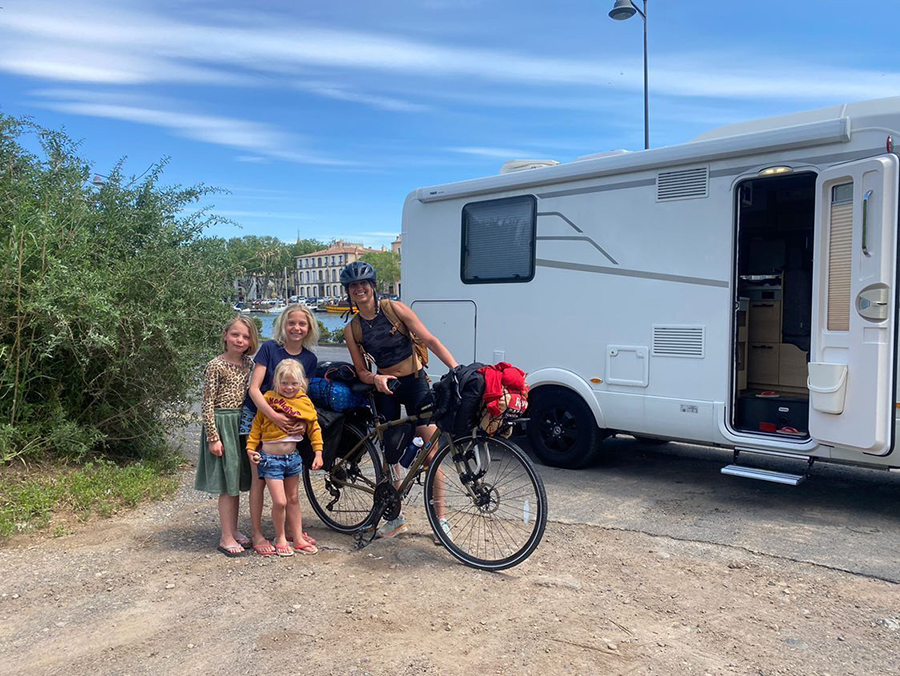 Gina stands with three blond children and her bike outside of a camper.