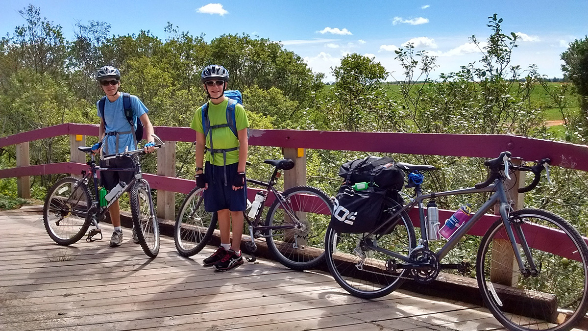 Sarah's two sons, white with brown hair, smile as they stand next to their mountain bikes on a bridge.