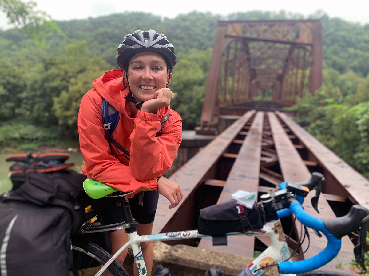 Sydney stands with her bike in rural Kentucky next to a derelict train bridge, a huge smile on her face.