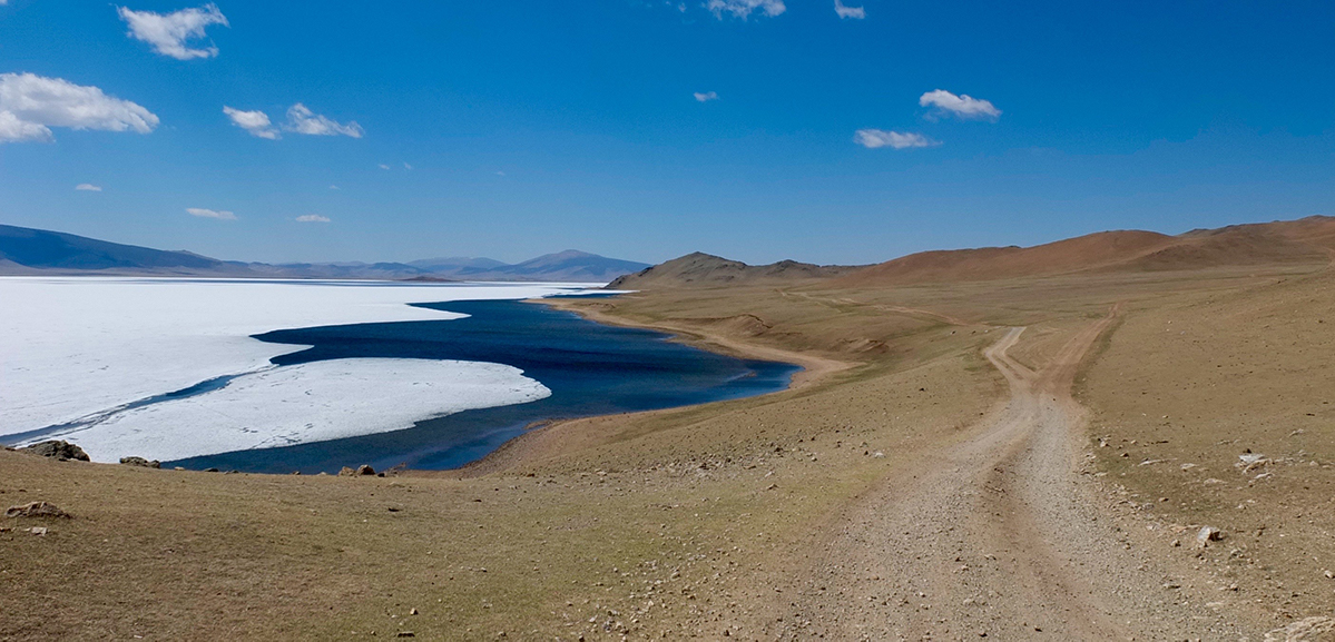 A dirt road snakes along a lakeshore, the water covered in ice and snow