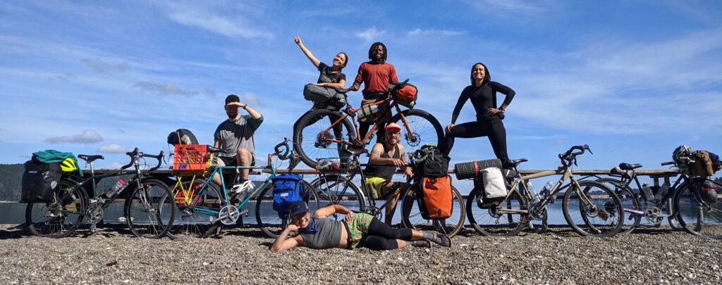 A group of smiling people with their bikes around and on top of a picnic bench.