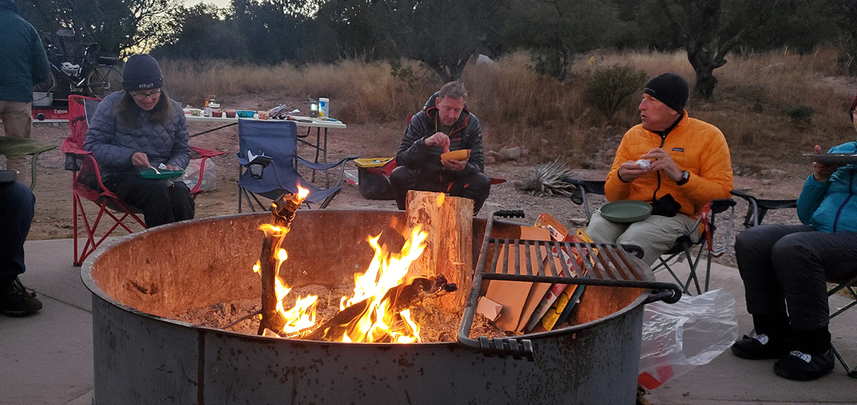 People chewing and talking around a metal fire pit.