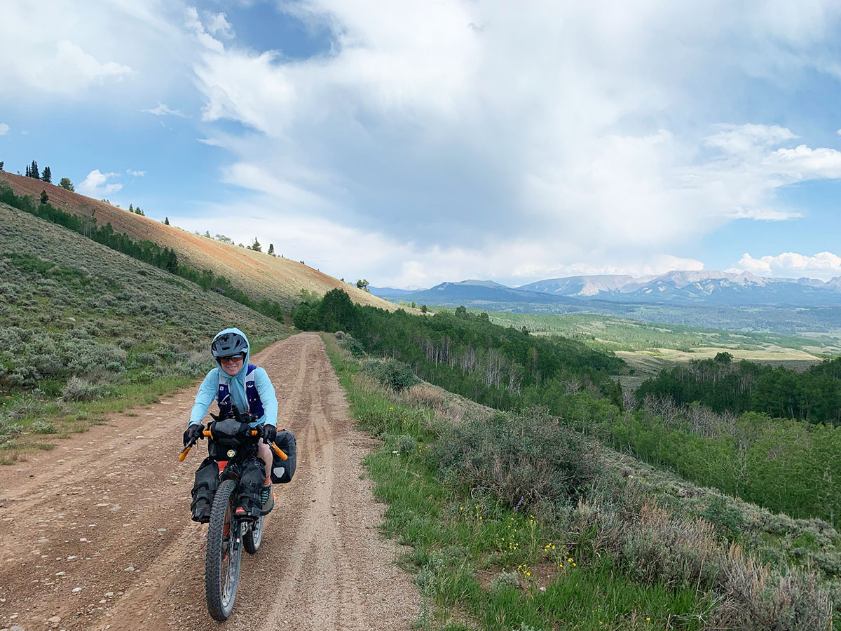 Irena or Sarah coasts along a dirt road in mountainous Montana. A big open view with dark thunderclouds is in the background.