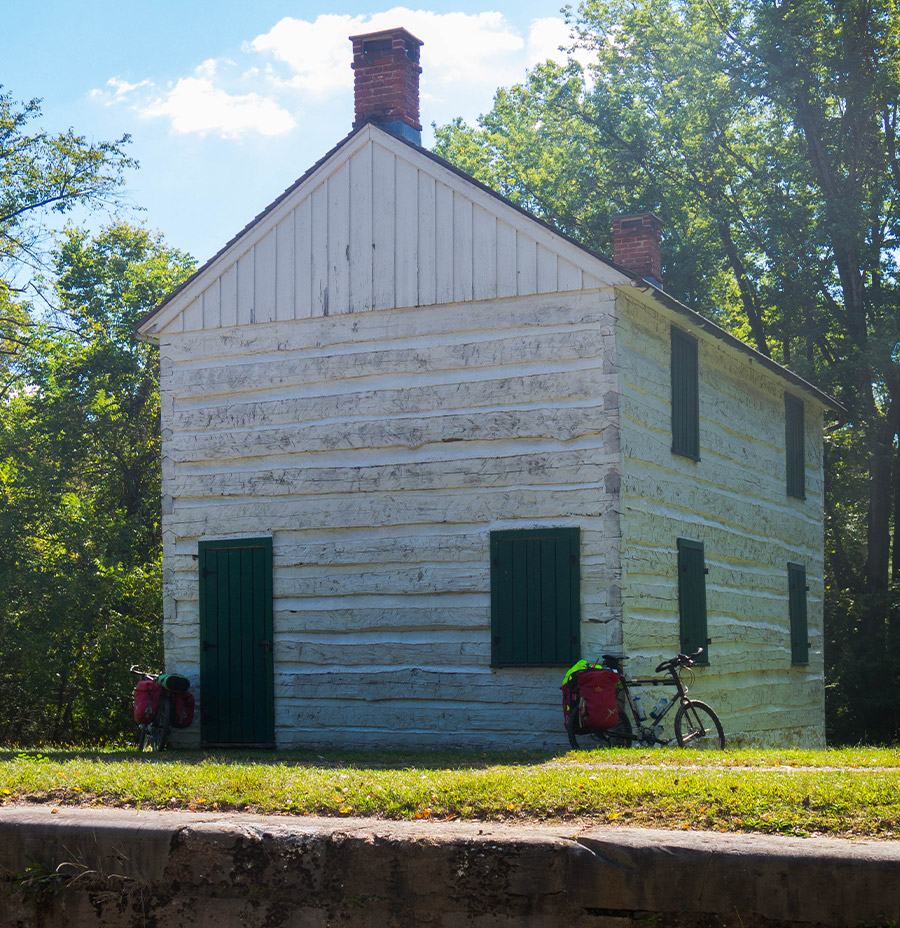 A bike leans against a tall, old white building. The rest of the scene is rural with green trees and grass.