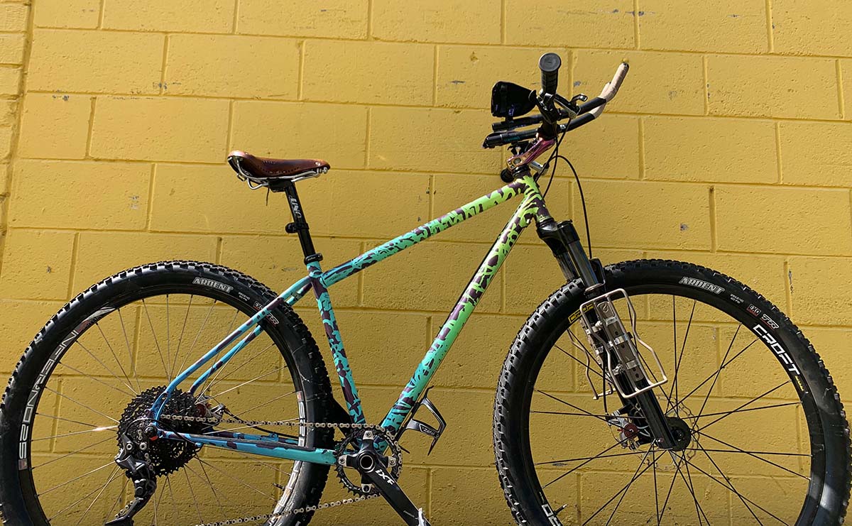 A splatter painted steel mountain bike rests against a bright yellow wall.