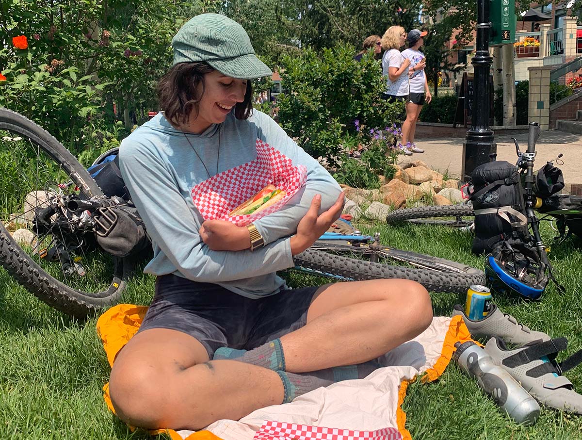 Mia cradles a sandwich in a basket in her arms while sitting in the grass in a park.