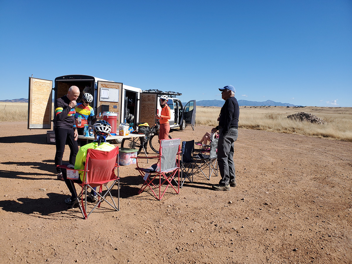 People sitting on camp chairs in the desert. Others getting snacks from a van with a trailer.
