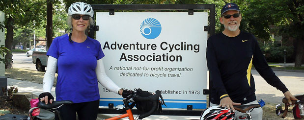 A man and a woman stand beside the Adventure Cycling Association sign in Missoula, MT, with their loaded bicycles.