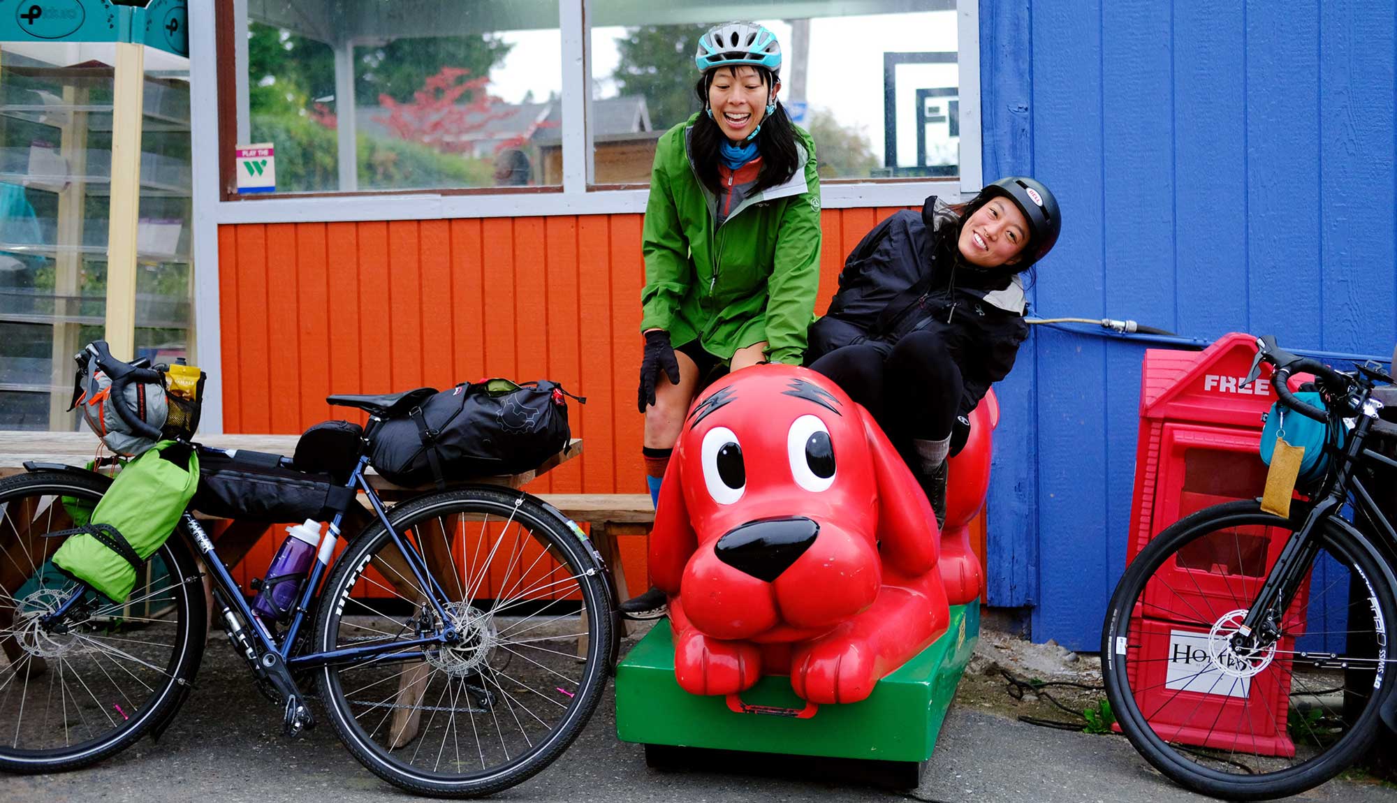 Two people in bike helmets grinning and riding a Clifford the Big Red Dog child's ride.